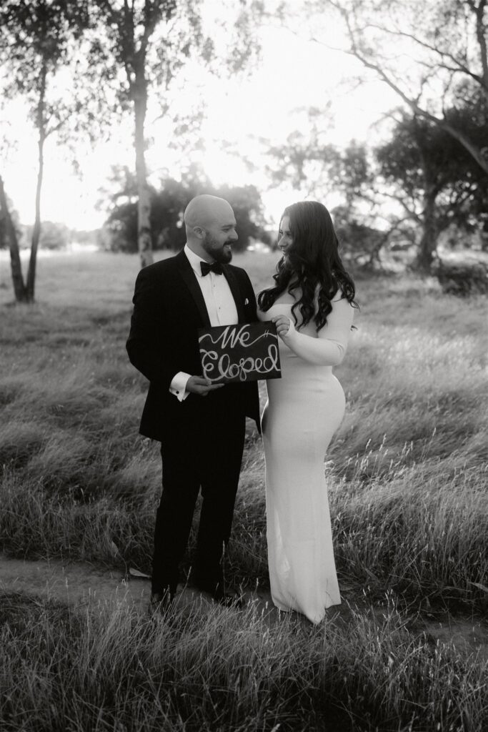 A couple in wedding attire holding a sign to announce their surprise elopement